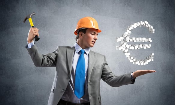Businessman going to crash with hammer euro currency sign. Young handsome man in business suit and safety helmet standing on grey wall background. Financial crisis, bankruptcy and inflation pressure.