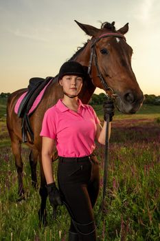 Beautiful smiling girl jockey stand next to her brown horse wearing special uniform on a sky and green field background on a sunset. Equitation sport competition and activity.