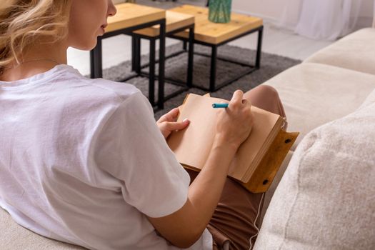 Close up woman hand writing on notebook. Young woman sitting on sofa in modern apartment, casual style indoor shoot