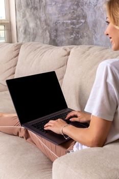 Woman on sofa working on laptop with mockup blank screen. Empty copy space on monitor for advertisement, language learning ad, online shopping website, social media site. Black screen.