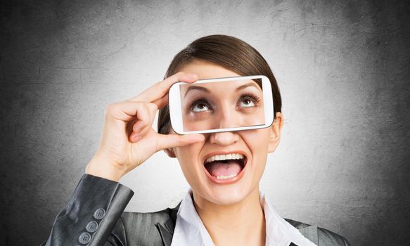 Portrait of pretty woman covering her eyes with smartphone. Happy businesswoman showing mobile phone with eyes on screen. Corporate businessperson on grey wall background. Mobile communication layout.