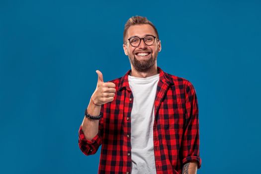 Emotional and people concept: young bearded man in checkered shirt. Hipster style. Studio shot on blue background. Copy space. The guy shows his hand - It's okay