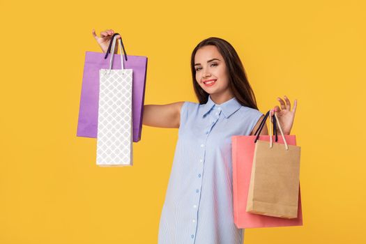 Image of a young brunette lady in white and blue summer striped dress posing with shopping bags and looking at camera over yellow background. Shopping. Sale
