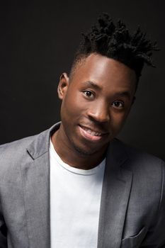 Close-up portrait of handsome black man with charming smile. Studio shot of well-dressed african guy wears white T-shirt and gray jacket, on dark background
