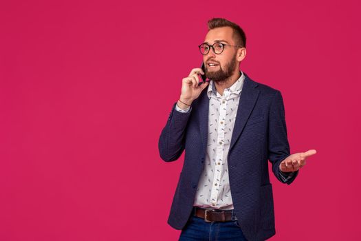 Young cheerful lawyer or businessman is standing on the pink background and smiling,talking on the phone, wearing jacket, shirt and jeans. Copy space