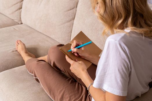 Close up woman hand writing on notebook. Young woman sitting on sofa in modern apartment, casual style indoor shoot