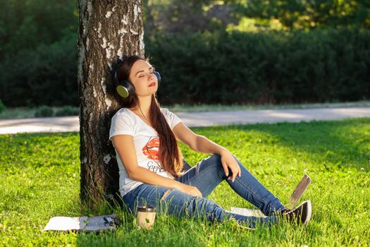 Woman relax with headphones listening to music sitting on grass in park. Young woman enjoys music and loneliness