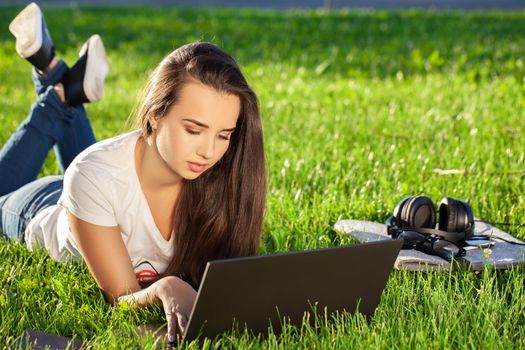 Young woman using laptop in the park lying on the green grass. Modern lifestyle, information technology or online shopping concept