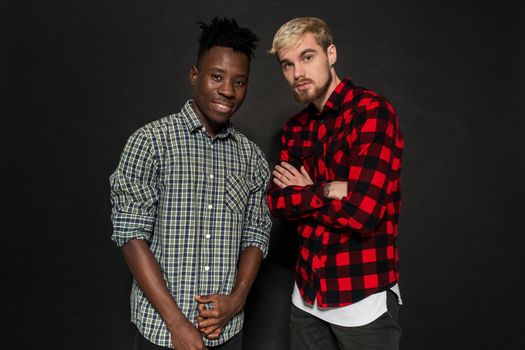 Studio shot of two stylish young men having fun. Handsome bearded hipster in a shirt in a cage standing next to his African-American friend against a dark background. International friendship concept.