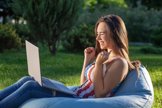 Young cute girl with laptop, sit on bean bag in garden or park, on green grass. Success small business, modern lifestyle, information technology, or online shopping concept