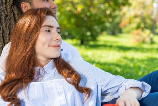 Young couple relaxing under tree in summer park on sunny day. Happy couple in love spend time outdoors together. Handsome man and pretty redhead girl enjoying each other. Romantic relationships.
