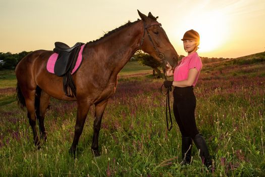 Beautiful smiling girl jockey stand next to her brown horse wearing special uniform on a sky and green field background on a sunset. Equitation sport competition and activity.