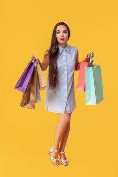 Image of a young brunette lady in white and blue summer striped dress posing with shopping bags and looking at camera over yellow background. Shopping. Sale