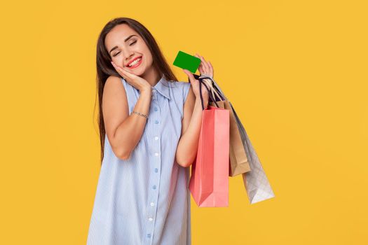 Trendy woman 20s in dress with long brown hair smiling while holding different shopping packages and card in hands isolated over yellow background. Shopping. Sale