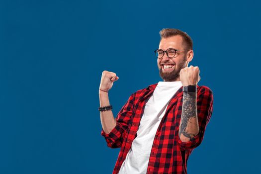 Emotional and people concept: young bearded man in checkered shirt. Hipster style. Studio shot on blue background. Copy space. Winner