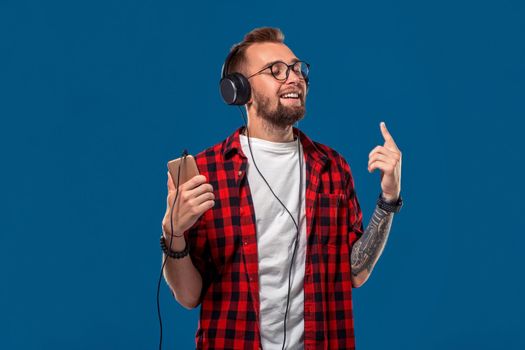 Dance Handsome young stylish man in headphones holding MP3 Player and dancing while standing against blue background. Emotional and people concept: young bearded man in checkered shirt. Hipster style. Copy space