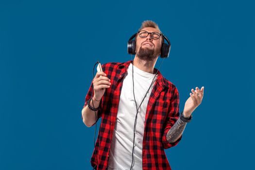 Happy young man listening to music with headphones. Handsome smiling guy in checkered shirt with closed eyes dancing with headphones. Isolated on blue background. Concept of people emotions
