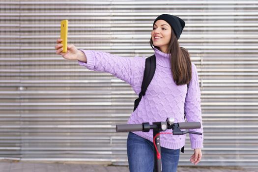 Woman in her twenties with electric scooter taking a selfie with a smartphone outdoors. Lifestyle concept.