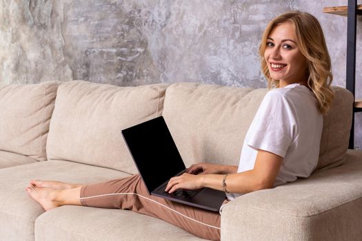 Woman on sofa working on laptop with mockup blank screen. Empty copy space on monitor for advertisement, language learning ad, online shopping website, social media site. Black screen.