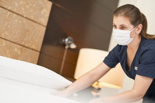 Young maid in mask looking at the bed and cleaning the room. Hotel service concept