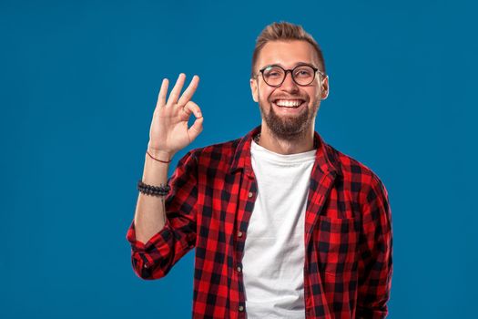 Emotional and people concept: young bearded man in checkered shirt. Hipster style. Studio shot on blue background. Copy space. The guy shows his hand - It's okay or OK.