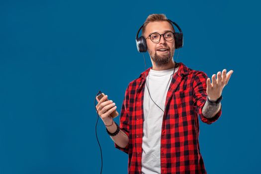 Happy young man listening to music with headphones. Handsome smiling guy in checkered shirt dancing with headphones. Isolated on blue background. Concept of people emotions