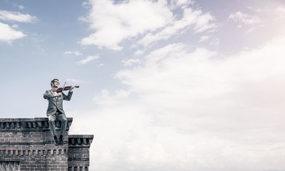 Young man wearing suit and glasses sitting on roof and playing violin