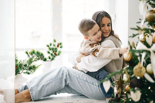 Happy mother with son sit on windowsill near Christmas tree, portrait