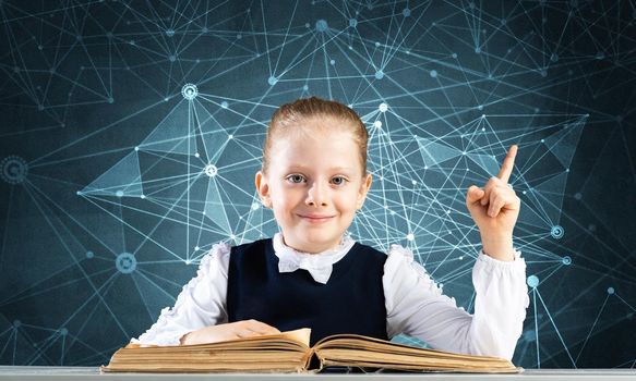 Smiling little girl sitting at desk with open book. Research and education in school. Happy schoolgirl in school wear finger pointing up. Clever girl reading book on deep blue plexus background