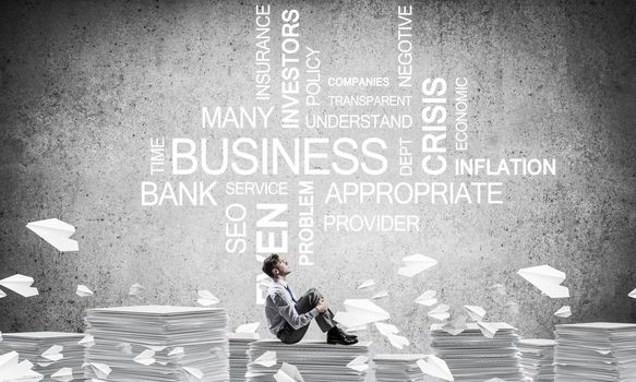 Businessman looking away while sitting on pile of documents among flying paper planes with business-related terms on background. Mixed media.