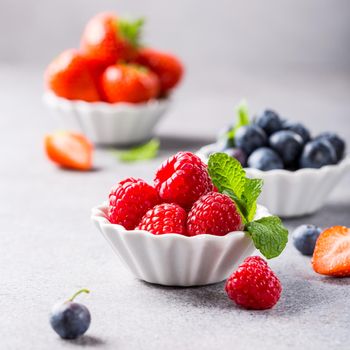 Fresh ripe raspberries, strawberries and bluebetties in white bowls on light gray concrete background. Healthy food concept with copy space.