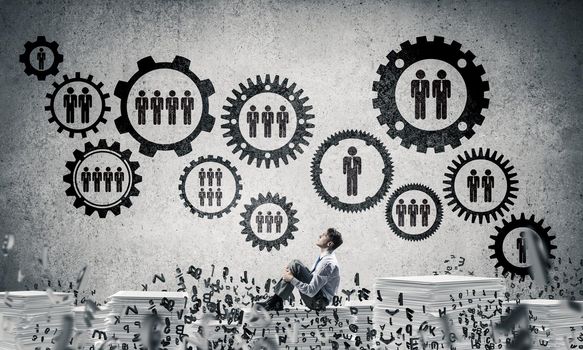 Businessman looking away while sitting on pile of documents among flying letters with social gear structure on background. Mixed media.