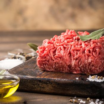 Fresh raw beef minced meat with salt, pepper, olive oil and laurel leaves on dark wooden board. Healthy food concept with copy space.