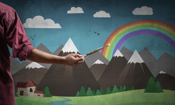 Close up artist hand holding paintbrush. Painter in shirt standing on background colorful picture. Summer landscape with mountains, blue sky and rainbow artwork. Creative hobby and profession.