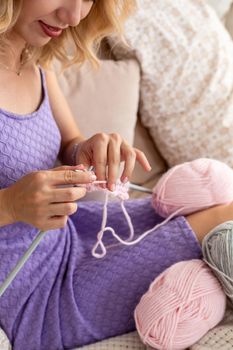 Close-up of woman in a lilac home dress knits a light pink scarf or plaid from natural threads on the bed. Close-up horizontal photo. Freelance creative working and living concept.