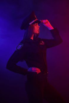 Pretty redheaded lady police officer in a uniform and a cap, with bright make-up is looking away and posing sideways against a black background with red and blue backlighting. Defender of citizens is ready to enforce a law and stop a crime.