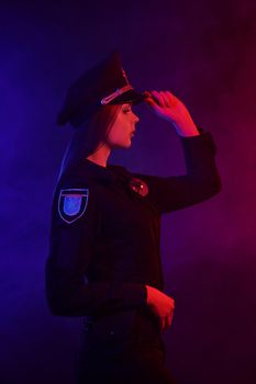 Pretty redheaded girl police officer in a uniform and a cap, with bright make-up is looking away and posing sideways against a black background with red and blue backlighting. Defender of citizens is ready to enforce a law and stop a crime.