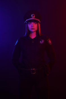 Young redheaded girl police officer in a uniform and a cap, with bright make-up is looking at the camera, has put her hand behind her back and posing against a black background with red and blue backlighting. Defender of citizens is ready to enforce a law and stop a crime.