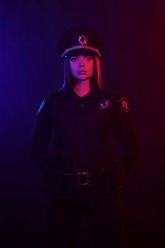 Young redheaded girl police officer in a uniform and a cap, with bright make-up is looking at the camera, has put her hand behind her back and posing against a black background with red and blue backlighting. Defender of citizens is ready to enforce a law and stop a crime.