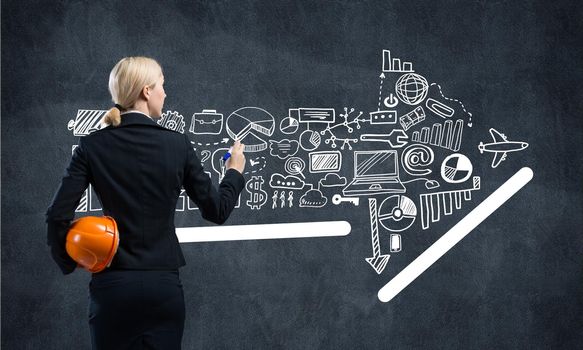 Businesswoman draws arrows consists of business doodles on blackboard. Back view woman with safety helmet and marker pen in hands. Idea generation and business development. Coaching and mentoring.