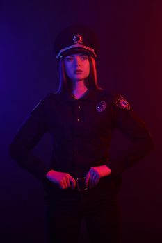 Young redheaded female police officer in a uniform and a cap, with bright make-up looking at the camera and posing against a black background with red and blue backlighting. Defender of citizens is ready to enforce a law and stop a crime.
