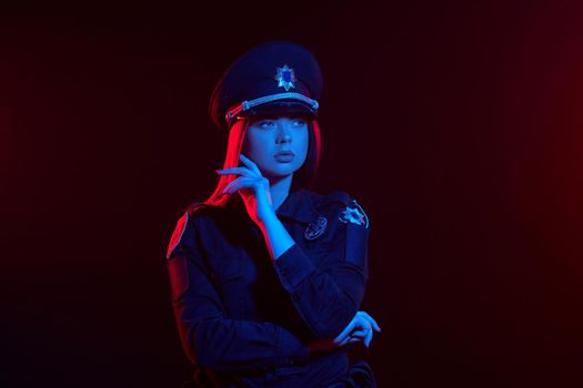 Close-up shot of an attractive redheaded woman police officer in a uniform and a cap looking away and posing with crossed hands against a black background with red and blue backlighting. Defender of citizens is ready to enforce a law and stop a crime.