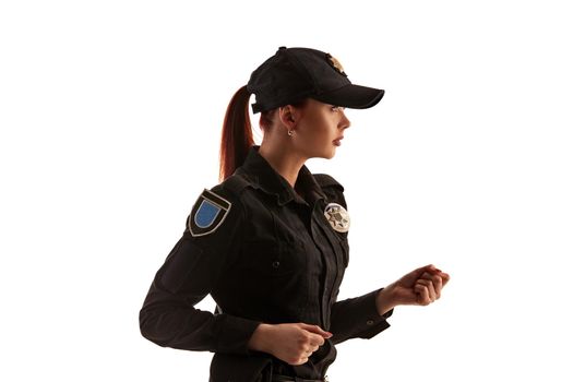 Close-up shot of a pretty redheaded policewoman in a black uniform and a cap preparing to fight, standing sideways isolated on white background. Defender of citizens is ready to enforce a law and stop a crime.