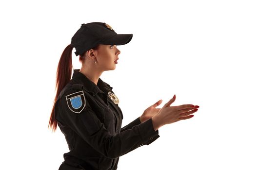 Close-up shot of a redheaded professional policewoman in a black uniform and a cap posing sideways and gesticulating, isolated on white background. Defender of citizens is ready to enforce a law and stop a crime.