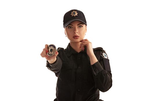 Close-up shot of an alluring redhead policewoman in a black uniform and a cap showing her badge and looking at the camera, isolated on white background. Defender of citizens is ready to enforce a law and stop a crime.