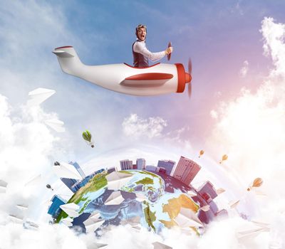 Man in aviator hat with goggles driving propeller plane. Earth globe with high modern buildings. Funny man having fun in small airplane. Blue cloudy sky with flying hot air balloons and paper planes