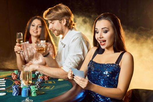Group of an elegant people playing poker at the gambling house. Focus on a emotional brunette in a blue shiny dress. Passion, cards, chips, alcohol, dice, gambling, casino - it is entertainment. Dangerous fun card game for money. Smoke background.
