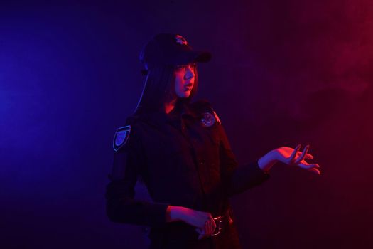 Beautiful redheaded maiden police officer in a uniform and a cap, with bright make-up is gesticulating while standing sideways against a black background with red and blue backlighting. Defender of citizens is ready to enforce a law and stop a crime.