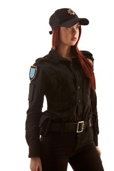 Serious redheaded policewoman in a black uniform and a cap is looking away and posing standing sideways, isolated on white background. Defender of citizens is ready to enforce a law and stop a crime.
