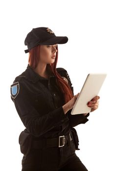 Pretty redhead policewoman in a black uniform and a cap is holding a tablet in her arms and typing something on it, isolated on white background. Defender of citizens is ready to enforce a law and stop a crime.